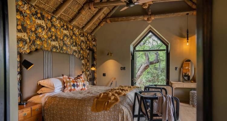 Chambre de style safari confortable avec toit de chaume, papier peint à motifs et vue sur une forêt verdoyante par une porte ouverte.