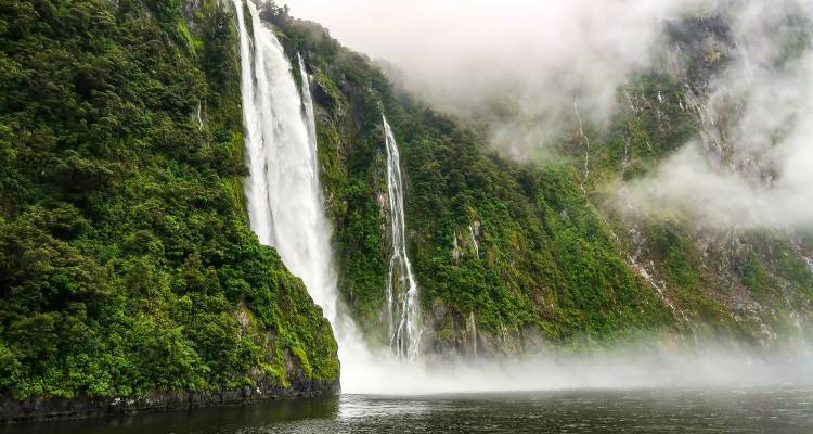 Tall waterfalls plunging down green cliffs into misty fiord waters.