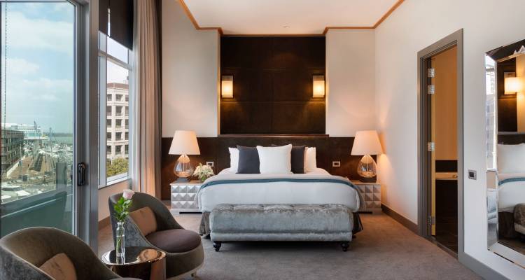 Luxurious city hotel room with king bed, plush bench and marina views through large windows.