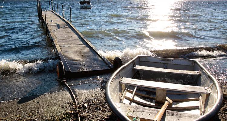 Small rowboat beached beside a wooden jetty on a sun-sparkled lake as gentle waves lap the shore.