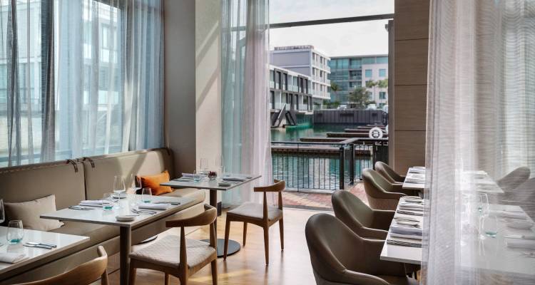 Modern restaurant with floor-to-ceiling sheer curtains, riverside terrace and neatly set tables for fine dining.