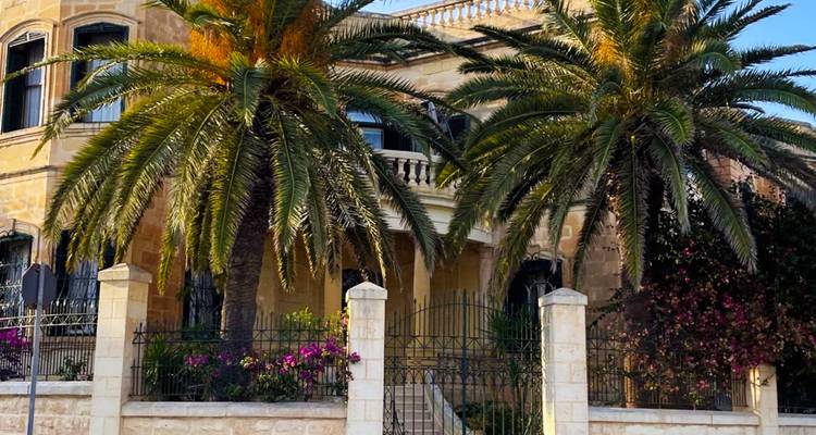 Elegant sandstone villa framed by tall palm trees and flowering shrubs behind an iron fence