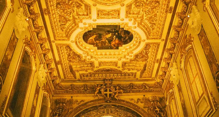 Richly ornamented gilded ceiling with central painting and Maltese cross emblem in a baroque hall