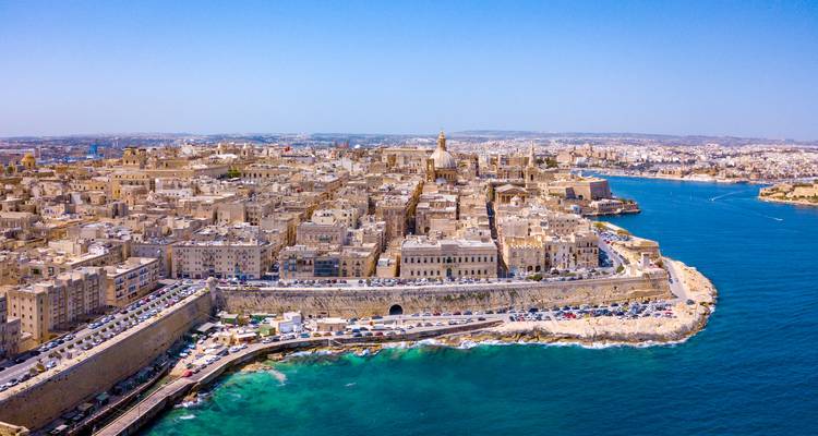 High aerial of Valletta peninsula showing dense sandstone architecture surrounded by vivid blue harbour waters