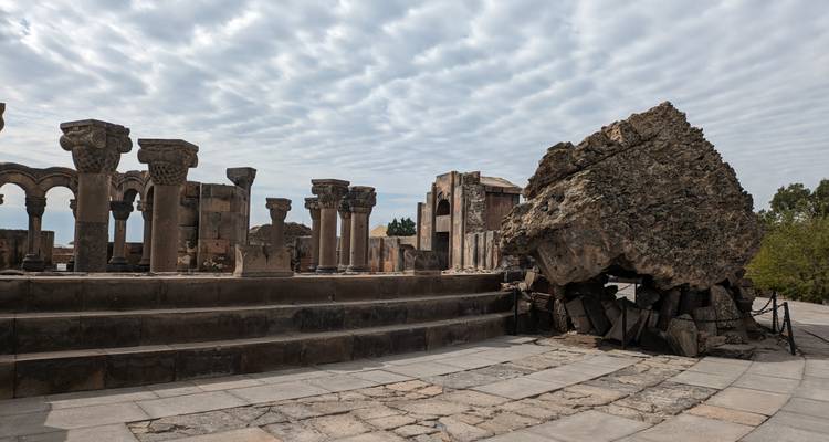 Row of basalt columns and toppled boulder at the circular ruins of an ancient cathedral under patterned clouds.
