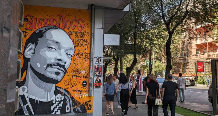 Urban street scene with colorful Snoop Dogg mural, boutique signs, and pedestrians strolling under shady trees.