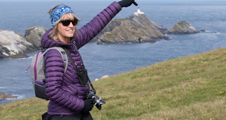 Une femme en tenue de randonnée pointe du doigt vers un phare sur une île à travers des falaises côtières ondulantes.