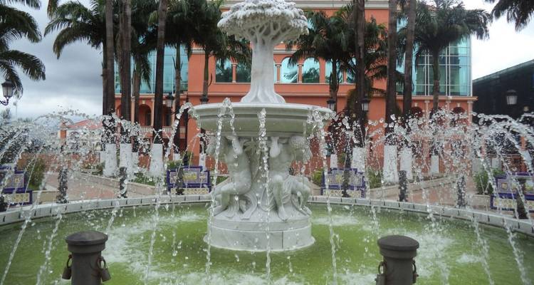 Close-up of an ornate white marble fountain surrounded by spurting water jets in a palm-lined plaza.