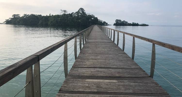 Wooden pier stretching far over calm tropical waters toward a small green island under a hazy sky.
