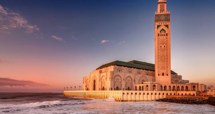 De Hassan II-moskee glint naast de Atlantische Oceaan terwijl golven uiteen spatten onder een roze zonsonderganghemel.
