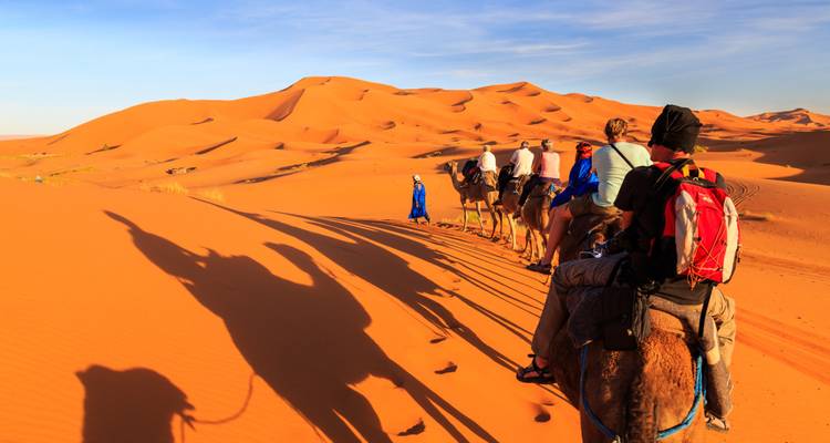Caravane de touristes chevauchant des chameaux à travers des dunes orange projetant de longues ombres à la lumière douce du soir