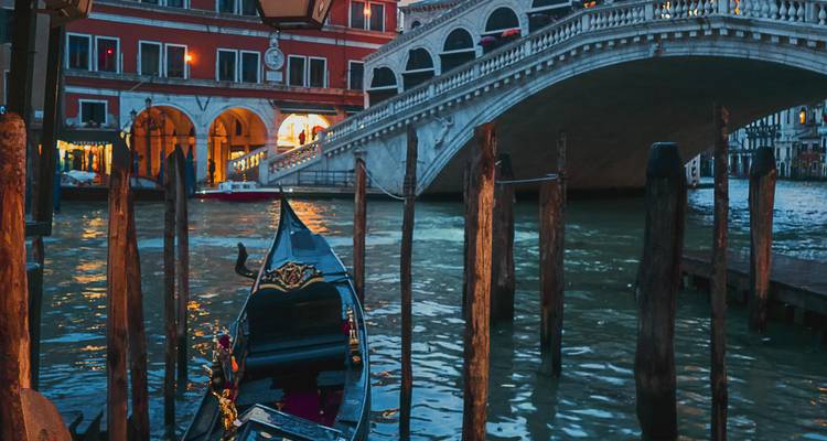 Moored gondola at dusk beside the illuminated Rialto Bridge spanning Venice's Grand Canal.