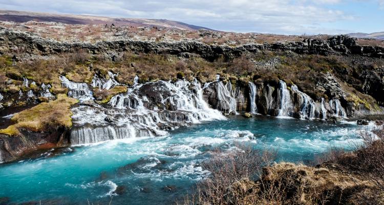 Turquoise glacial river cascading over multiple rocky ledges in a rugged Icelandic landscape.