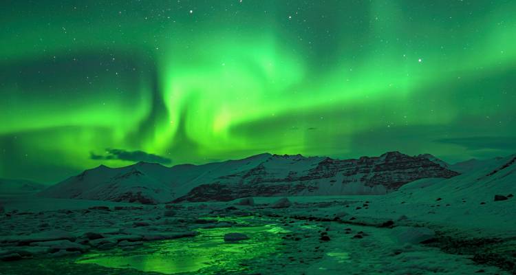 Brilliant green northern lights dancing over snow-covered mountains and icy river in Icelandic wilderness.