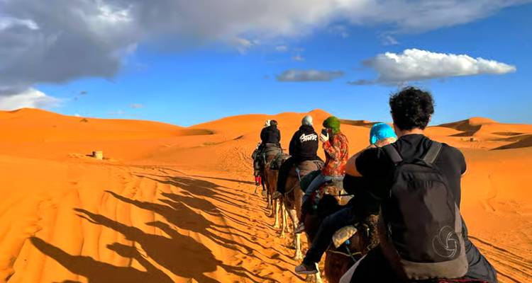 File de voyageurs à dos de chameaux traversant des dunes de sable orange vif avec des nuages épars au-dessus