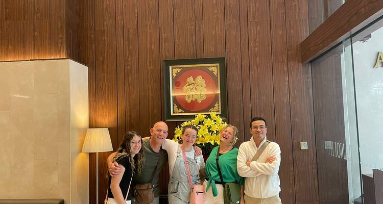 Five smiling travellers posing together in a hotel lobby with wooden panel walls and a yellow flower arrangement behind them