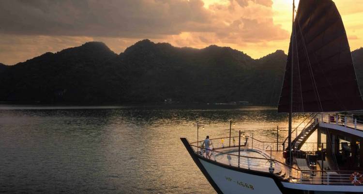Traditional junk-style cruise boat gliding past dark green karst hills at golden hour