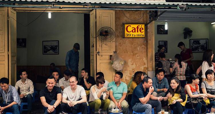 Busy local café front in narrow Vietnamese street with patrons seated on low stools beneath a yellow façade