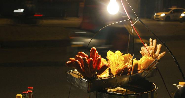 Night-time street food stall with sausages and fritters lit by a single hanging bulb
