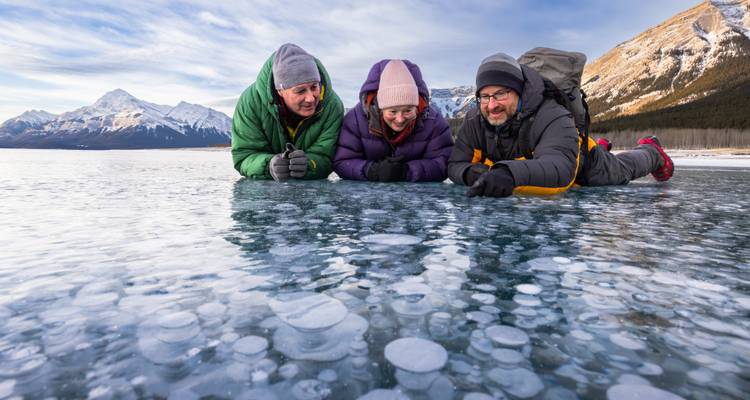 Trois voyageurs sont allongés sur un lac gelé cristallin parsemé de bulles de méthane, avec des pics de montagne enneigés s'élevant à l'arrière-plan.