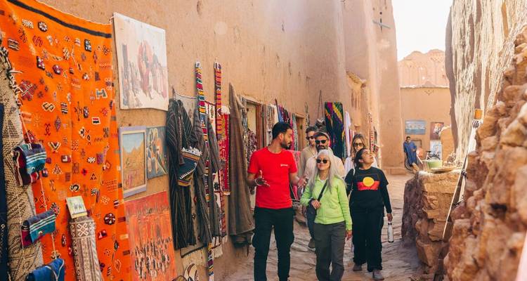 Travellers stroll a narrow clay alley lined with colourful carpets and crafts in Ait Benhaddou.