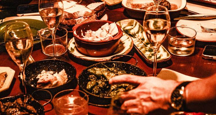 Warm, dimly lit table filled with tapas dishes, wine glasses and diners’ hands reaching for food.