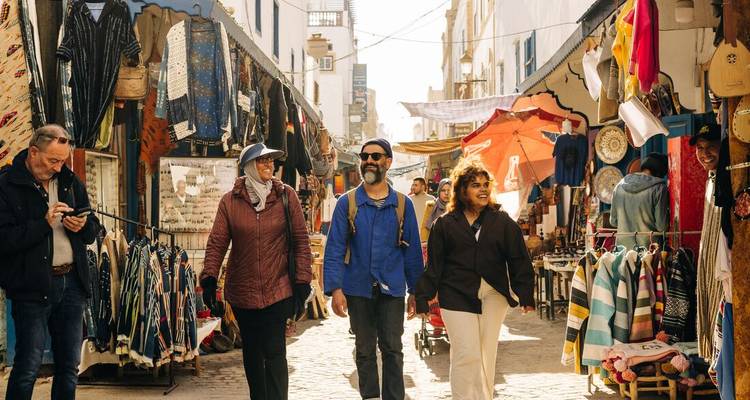 Tourists explore a lively lane of shops and textiles in Essaouira’s medina under warm sunlight.
