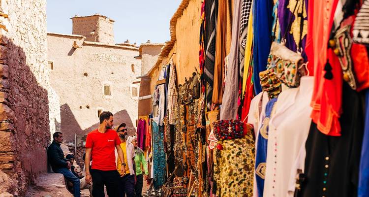 Visitors browse colourful hanging fabrics and souvenirs along an earthen alley in southern Morocco.