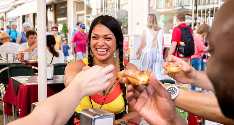 Friends laughing and sharing pastel de nata at an outdoor café on a sunny Lisbon street.