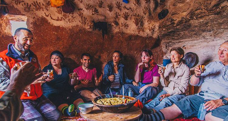 Un petit groupe assis à l'intérieur d'une maison berbère rustique partageant du thé et des aliments locaux, avec des empreintes de mains sur les murs en terre.