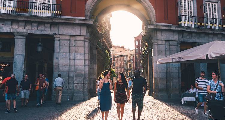Trois voyageurs marchent sous une arche de pierre illuminée par le soleil en direction du quartier historique de Madrid.