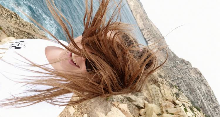 Close-up van een vrouw die lacht terwijl de wind haar haren blazen op zeerotsen.