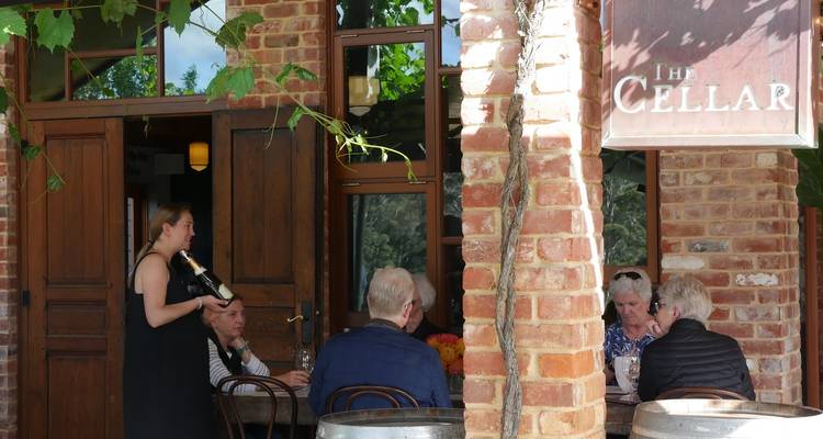 Des visiteurs assis à l'extérieur d'une porte de cave rustique en brique tandis qu'un serveur verse du vin à leur table.