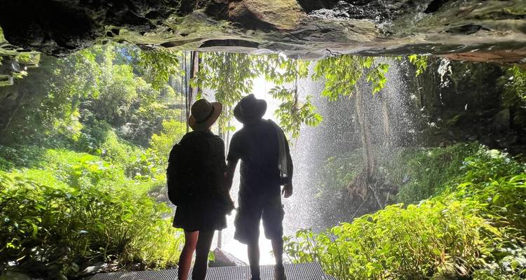 Una pareja silueteada dentro de la entrada de una cueva enmarcada por vegetación exuberante y una cascada alimentada por la lluvia.