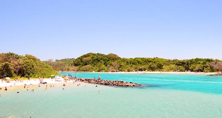 Bahía turquesa con nadadores y bañistas en una playa de arena blanca y brillante rodeada de bosque verde.