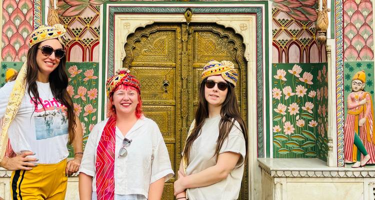Three women wearing colorful turbans pose before an ornate golden doorway adorned with lotus murals.