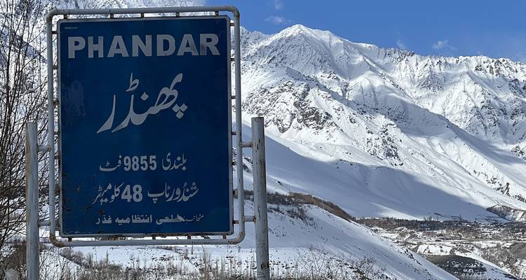 Blaues Straßenschild für Phandar vor dramatischen schneebedeckten Gipfeln und einem winterlichen Tal.