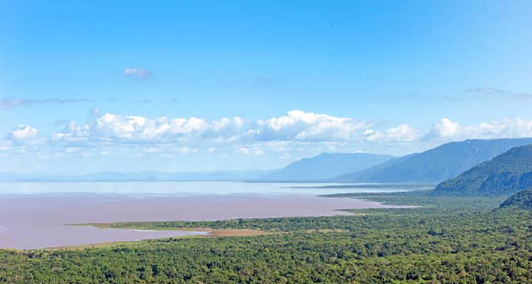 Wide view of a lake bordered by dense green forest and distant mountains under a bright blue sky.