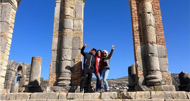 Trois voyageurs posant parmi les anciennes colonnes de pierre et les arcs de ruines romaines sous un ciel dégagé