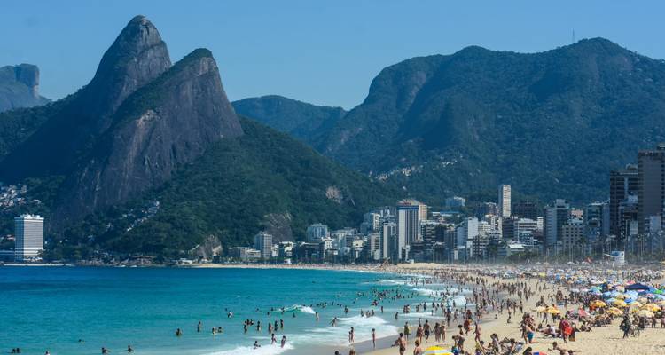 Plage d'Ipanema remplie de baigneurs, eau turquoise, gratte-ciel de la ville et les pics des Dois Irmãos dominant l'horizon.