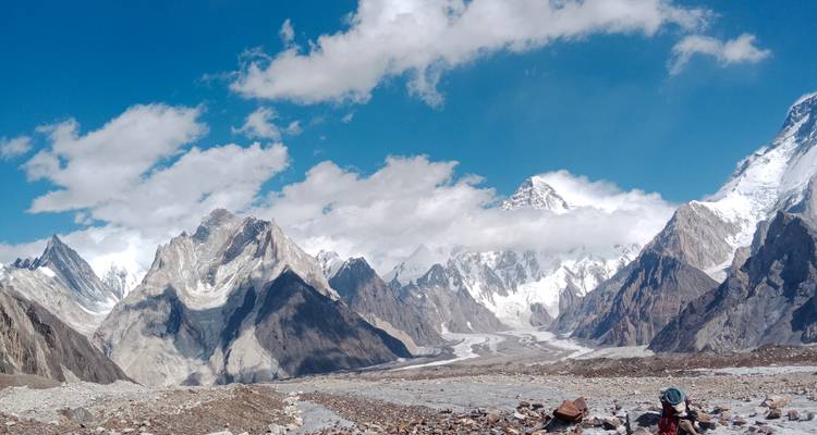 Large panorama d'une vallée glaciaire encadrée par des pics enneigés aux contours déchiquetés avec un randonneur solitaire marchant à travers une moraine rocheuse au premier plan.