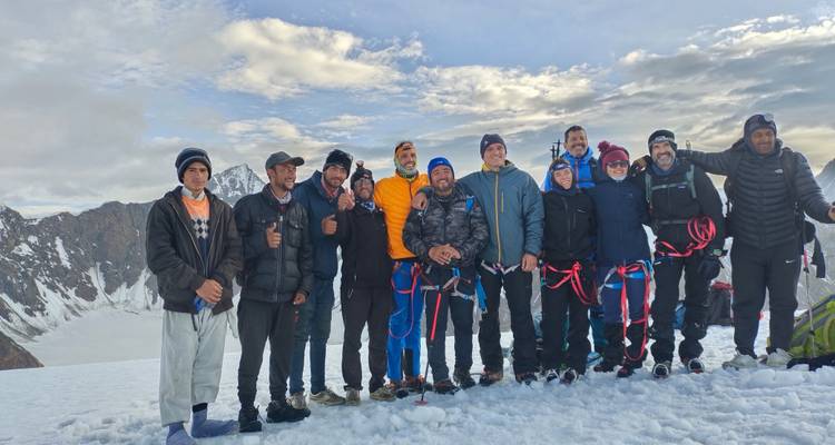 Groupe de trekking souriant posant sur un col enneigé avec des harnais d'escalade, entouré de paysages alpins élevés sous des nuages épars.