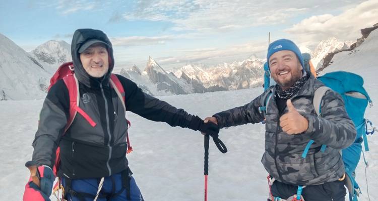 Deux alpinistes vêtus de tenues hivernales se serrent la main et sourient sur un plateau enneigé avec des pics dentelés en arrière-plan.