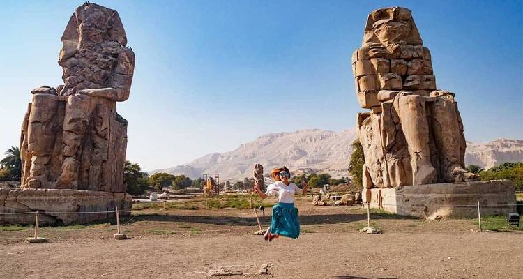 Traveler mid-jump between the massive Colossi of Memnon statues against a clear blue sky.