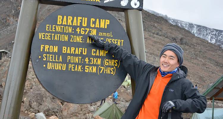 Traveller points proudly at circular Barafu Camp altitude sign on Kilimanjaro trail.