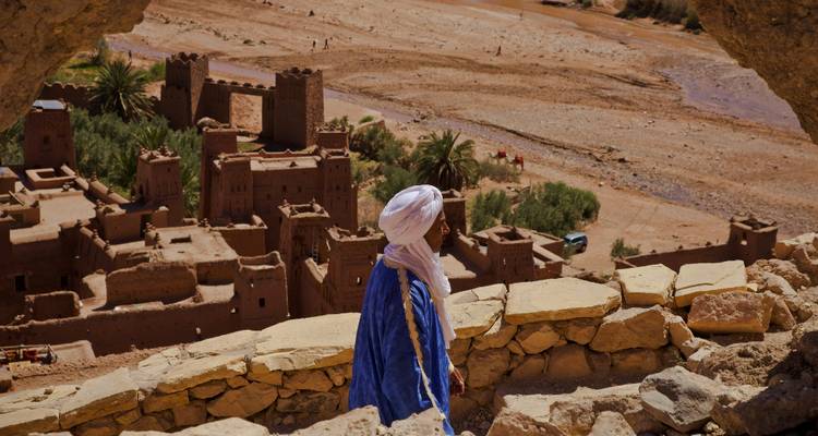 Un hombre bereber con una túnica azul está de pie en la cima de una kasbah de adobe, observando un valle de río desértico.
