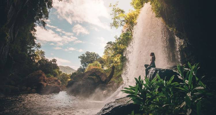 Traveler perched on rock beside misty waterfall cascading into rainforest pool under blue sky.