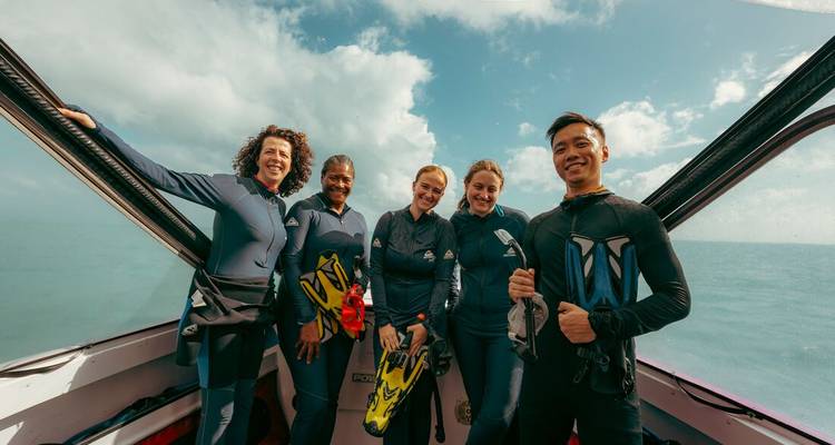 Smiling snorkelers in wetsuits pose on a boat deck holding fins and masks against an open sea backdrop.
