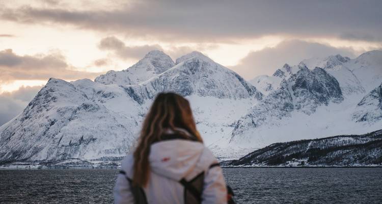 Woman in winter jacket looks out over dramatic snow-clad peaks at dusk by the sea.