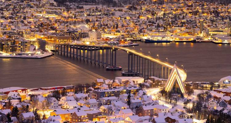 Night view of a snow-covered coastal city with the illuminated Tromsø Bridge and Arctic Cathedral reflecting in the water